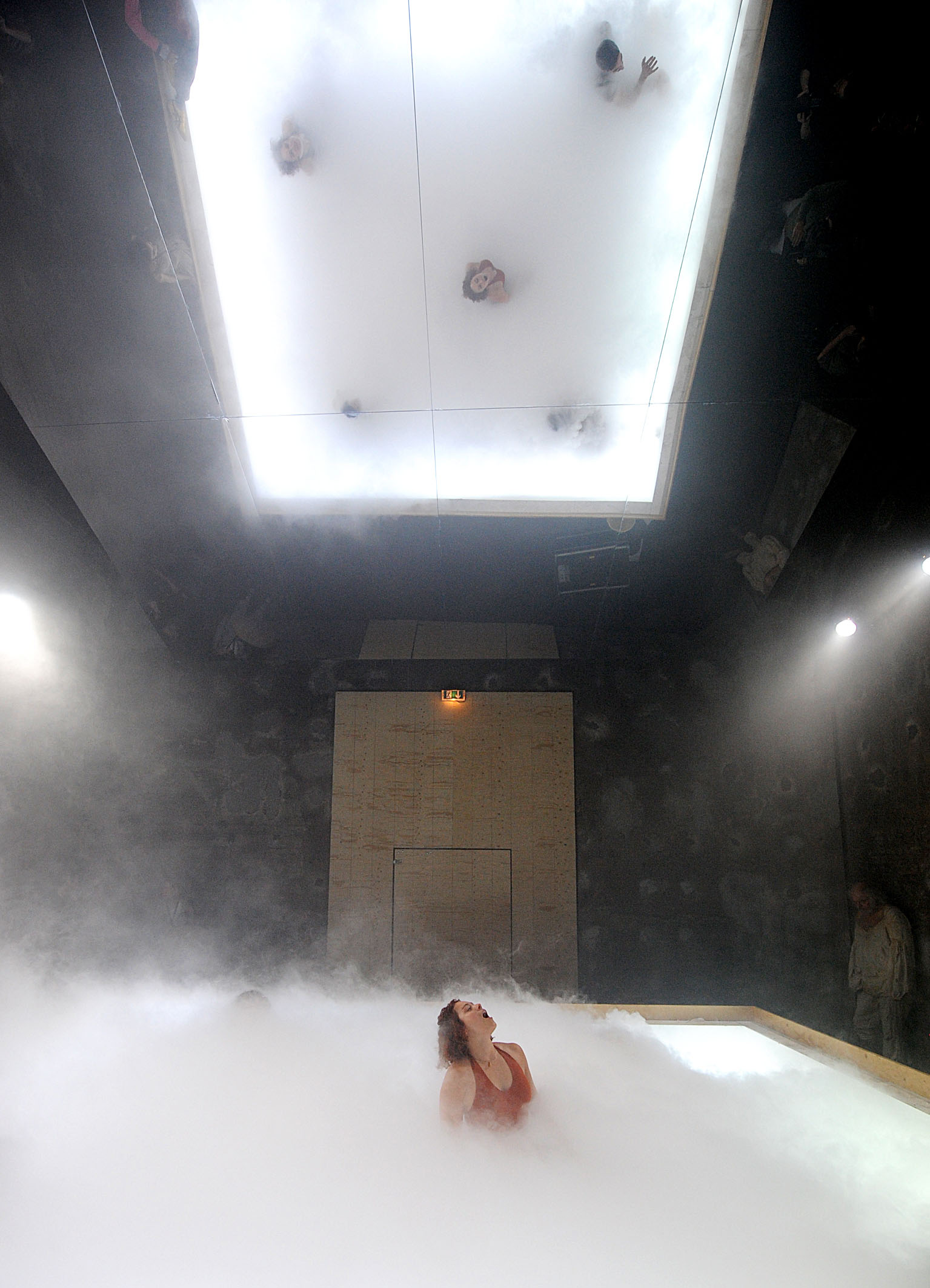 A large room with a mirror for a ceiling. A performer is seen emerging from a large box filled with dry ice and the heads of other performers appear in the reflection of the mirror.
