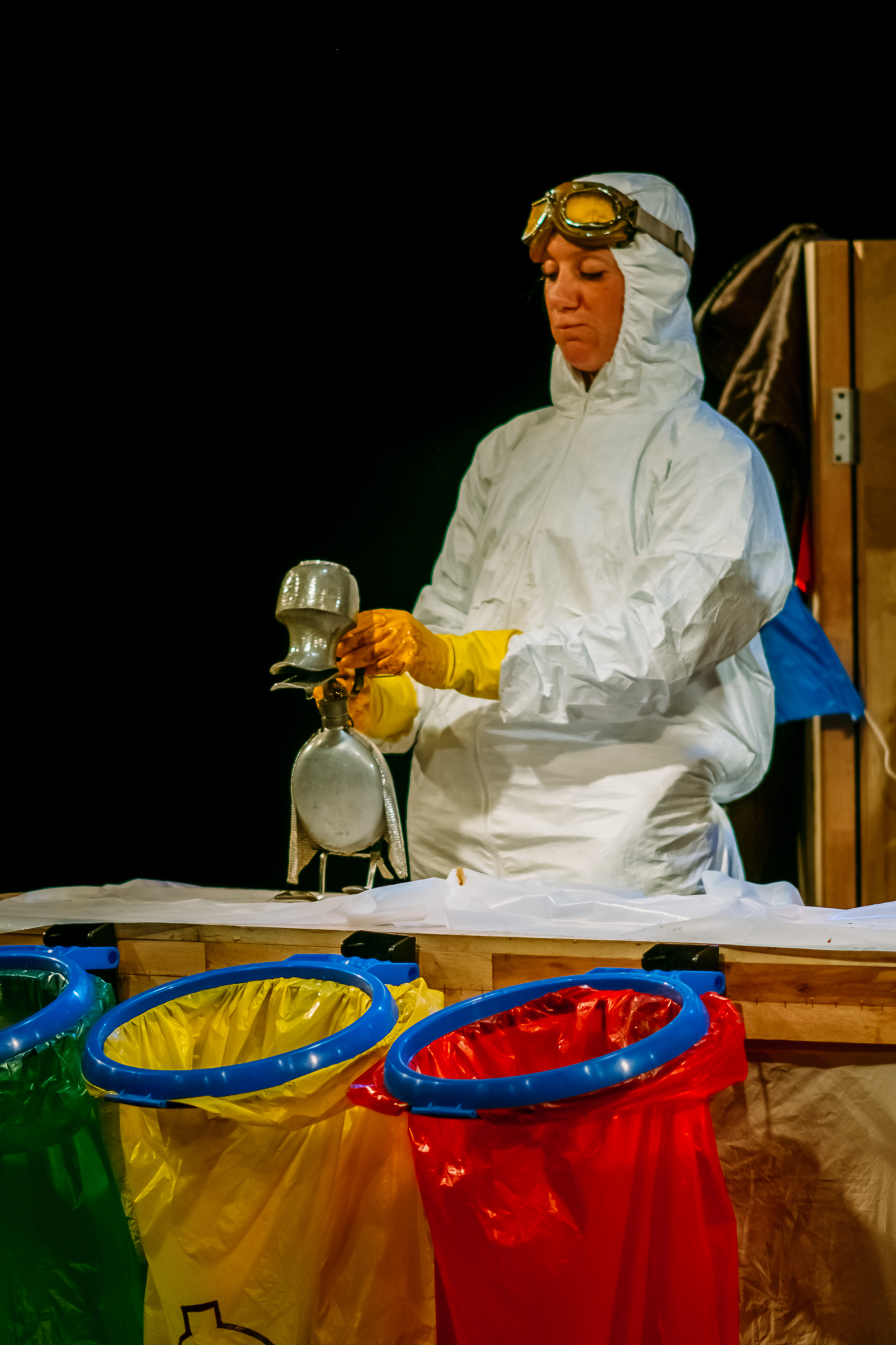 a performer dressed in white disposable boiler suit and yellow plastic gloves manipulates a puppet duck made from metal household items on a tabletop. A green, yellow and red plastic waste bag hang off the table in front of them.