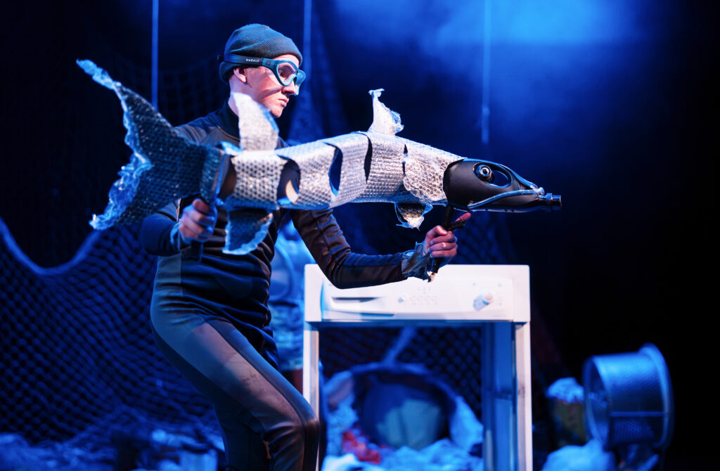 a puppeteer is animating a barracuda fish made out of black plastic bottles and bubble wrap. The fish is mid movement as if swimming in the sea.