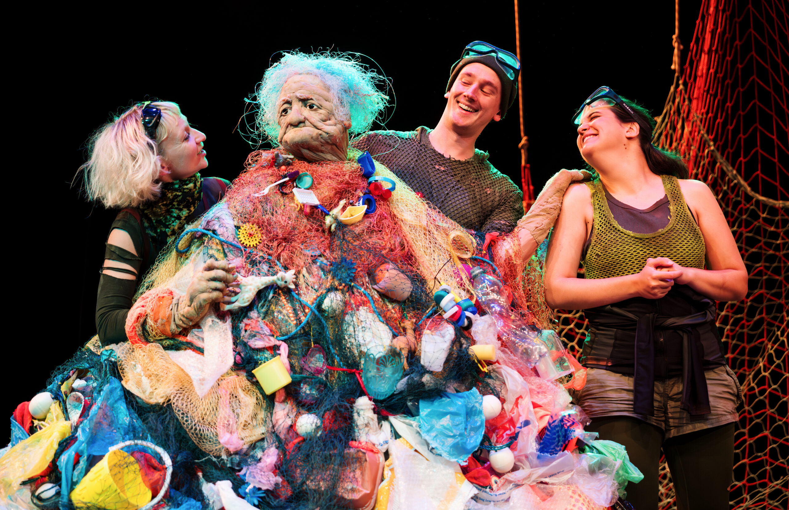 three performers laugh merrily with an ancient female puppet whose body is made out of plastic waste.