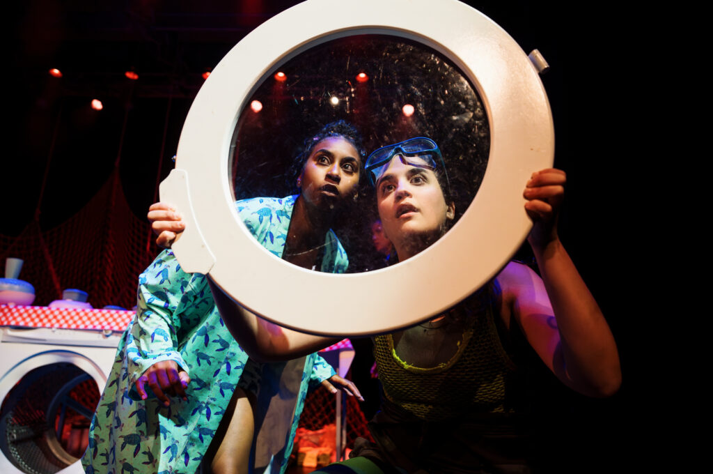 two performers stare through the glass door of a washing machine which has been removed from the machine. It looks as if they are staring out of the window of a space rocket.