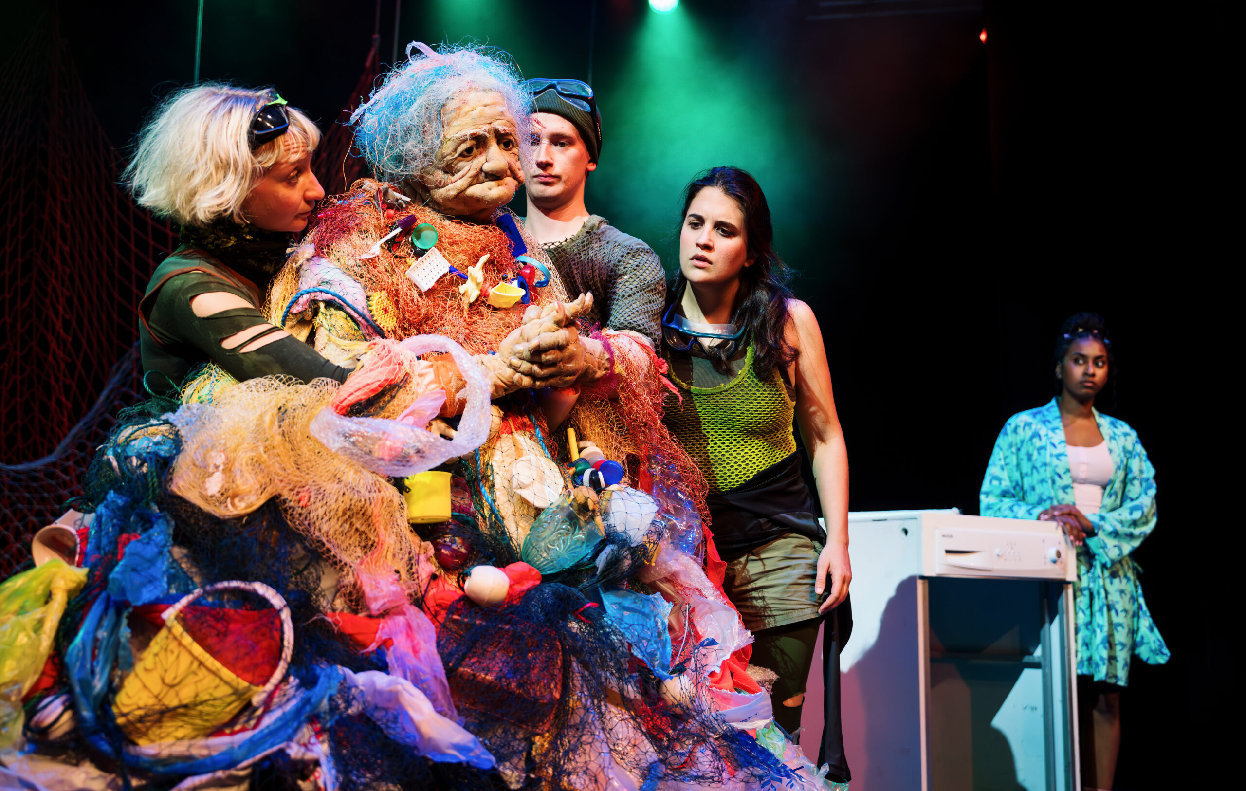 an ancient looking female spirit with a body made out of lots of plastic waste is being puppeteered by two puppeteers. A third performer is staring at the puppets hands and in the background a fourth performer is staring at them.