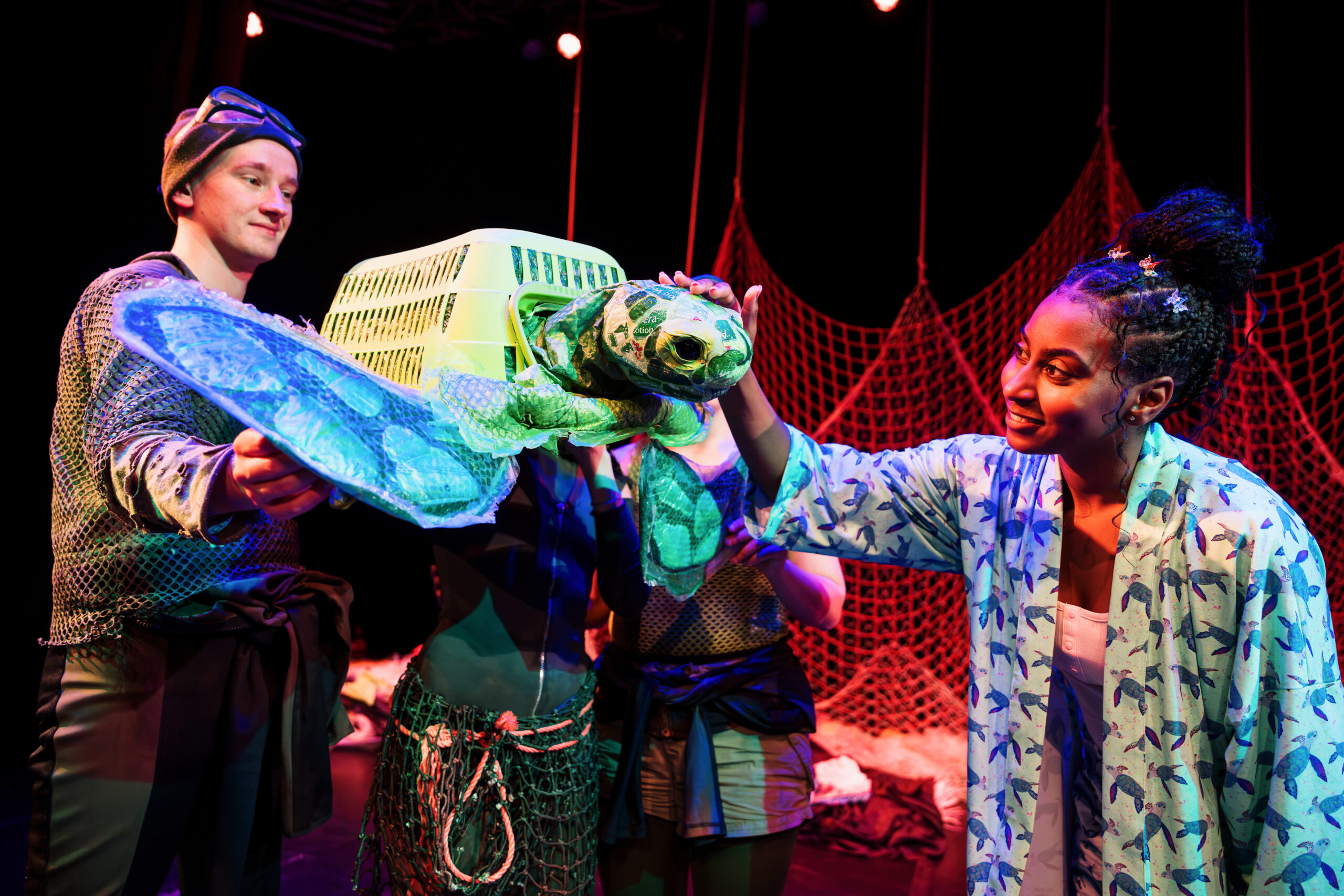 two puppeteers hold a puppet turtle made out of a green plastic washing basket wtih plastic head and fins. An actress in a dressing gown is stroking the turtles head and looking lovingly at it.