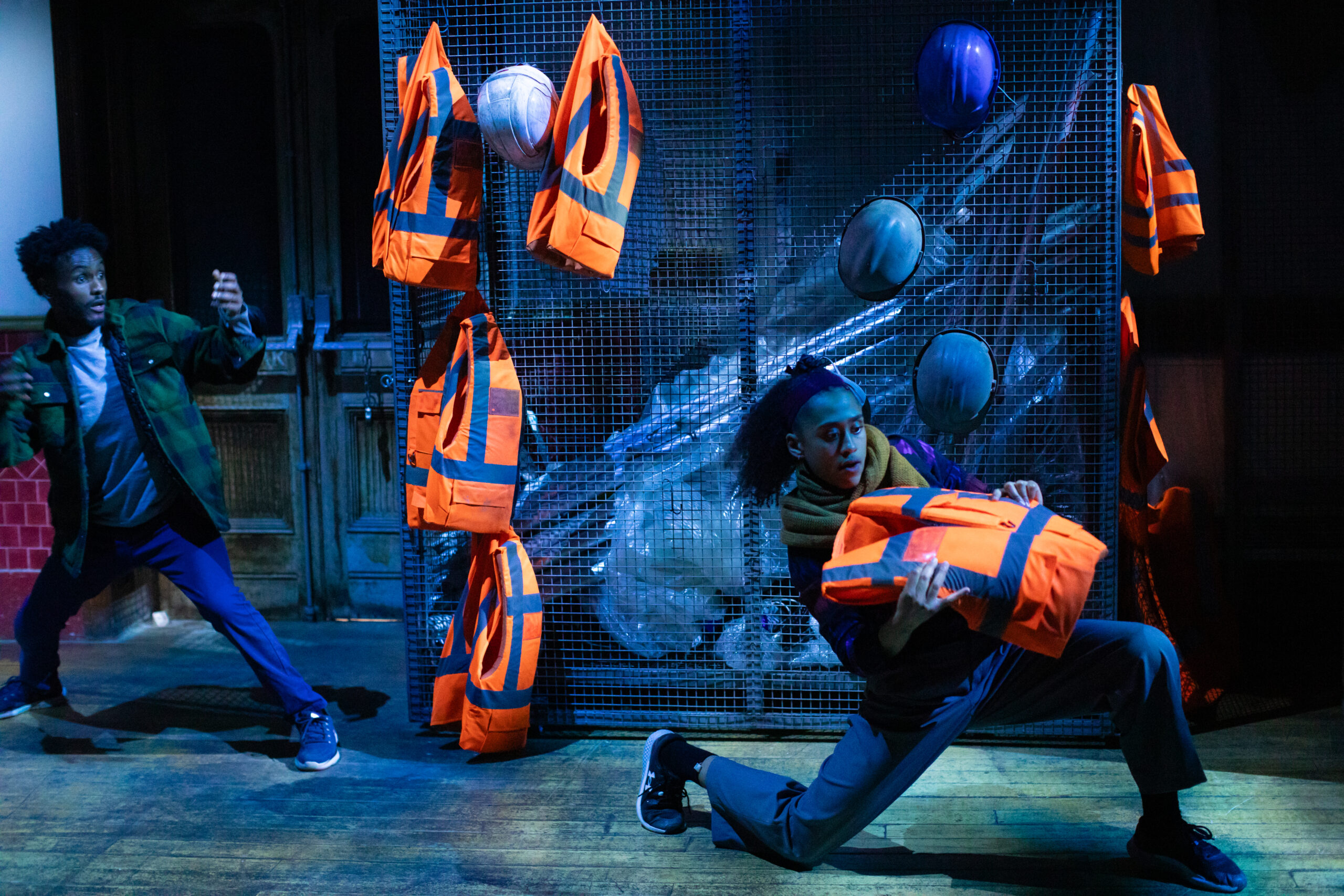 A closed cage in the background full of plastic. Hanging on the outside of the cage are hardhats and orange lifejackets. At the front of the image, a dancer is kneeling down and moving backwards holding one of the lifejackets. Behind her is another performer with arms at stretched, ready to catch a lifejacket, gently moving backwards.