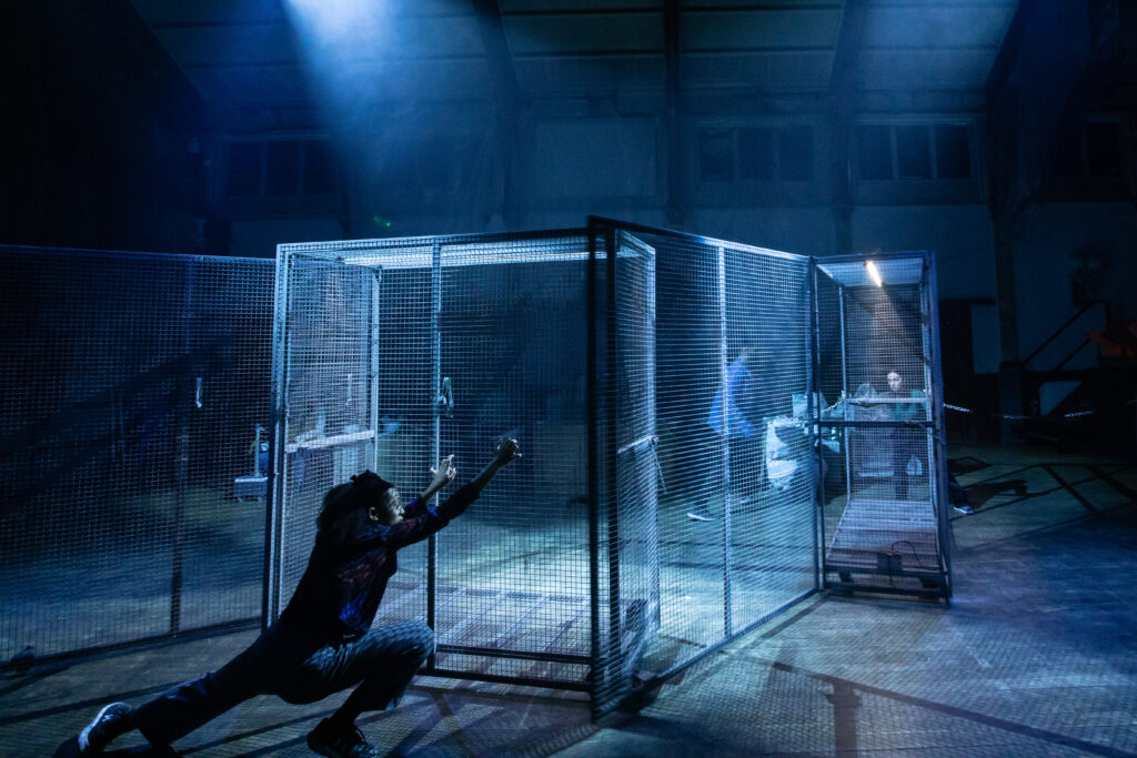Large open cages are connected to each other and look like a maze. the cages are on wheels and one performer is crouching down as if in movement, holding on to one side. In the distance, you can see two other performers moving through the space and one is also holding on another side of the cage.