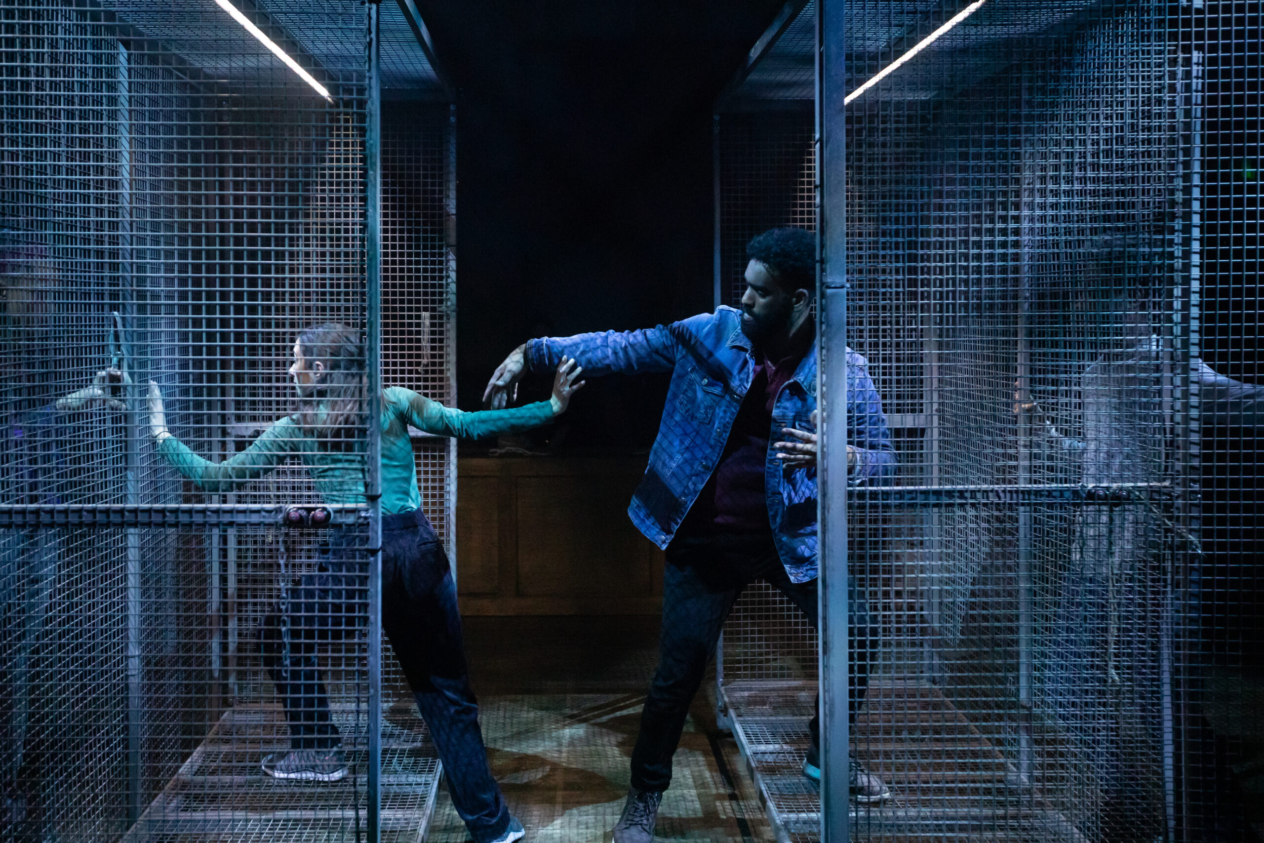 A female and male performer with their arms almost touching each with one foot inside two separate open cages. One performer is looking through her cage, and the other perfomer is looking at her.