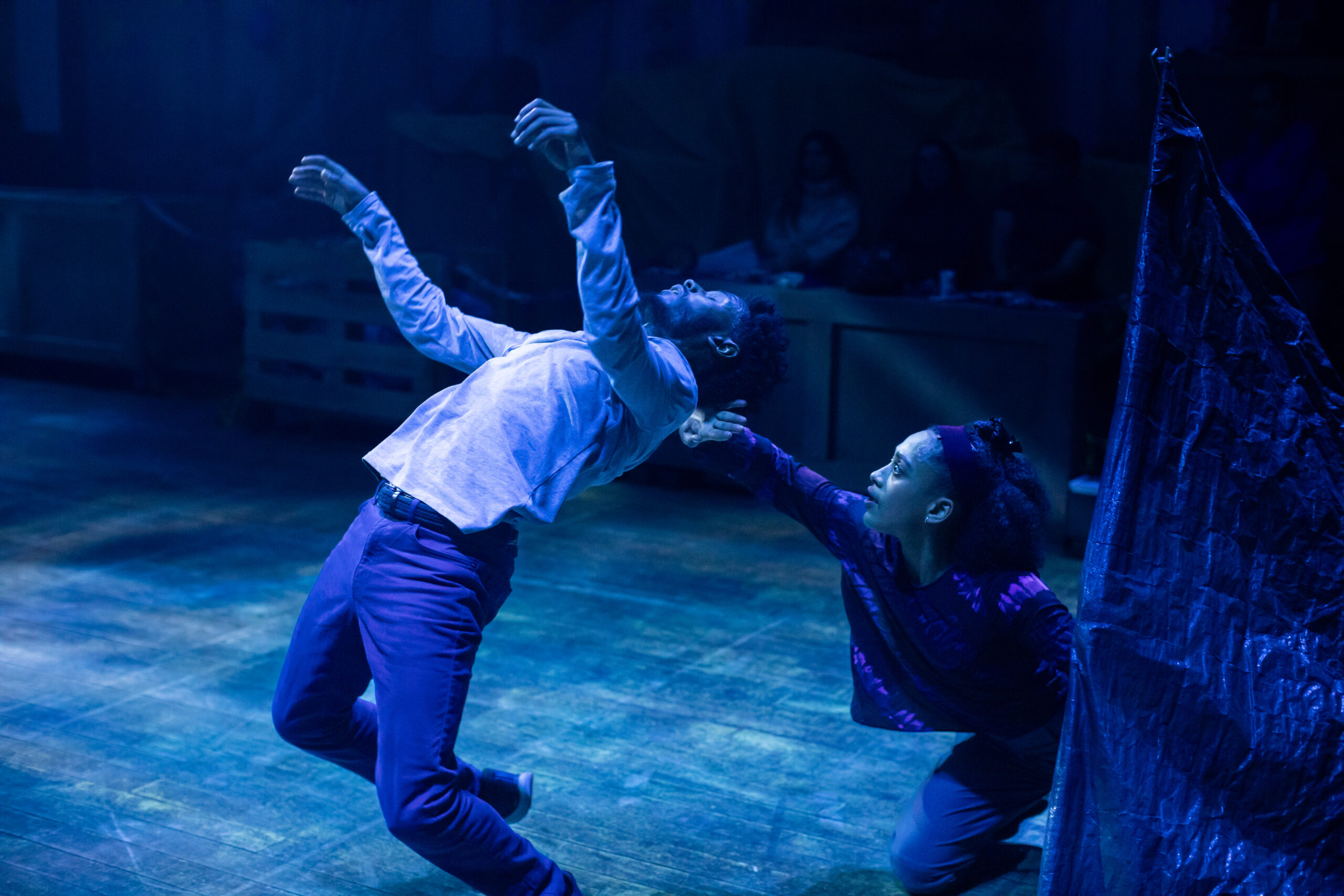 A dancer bends backwards bathed in blue light. His arms are outstretched, and behind him another dancer, kneeling on the ground is holding his head