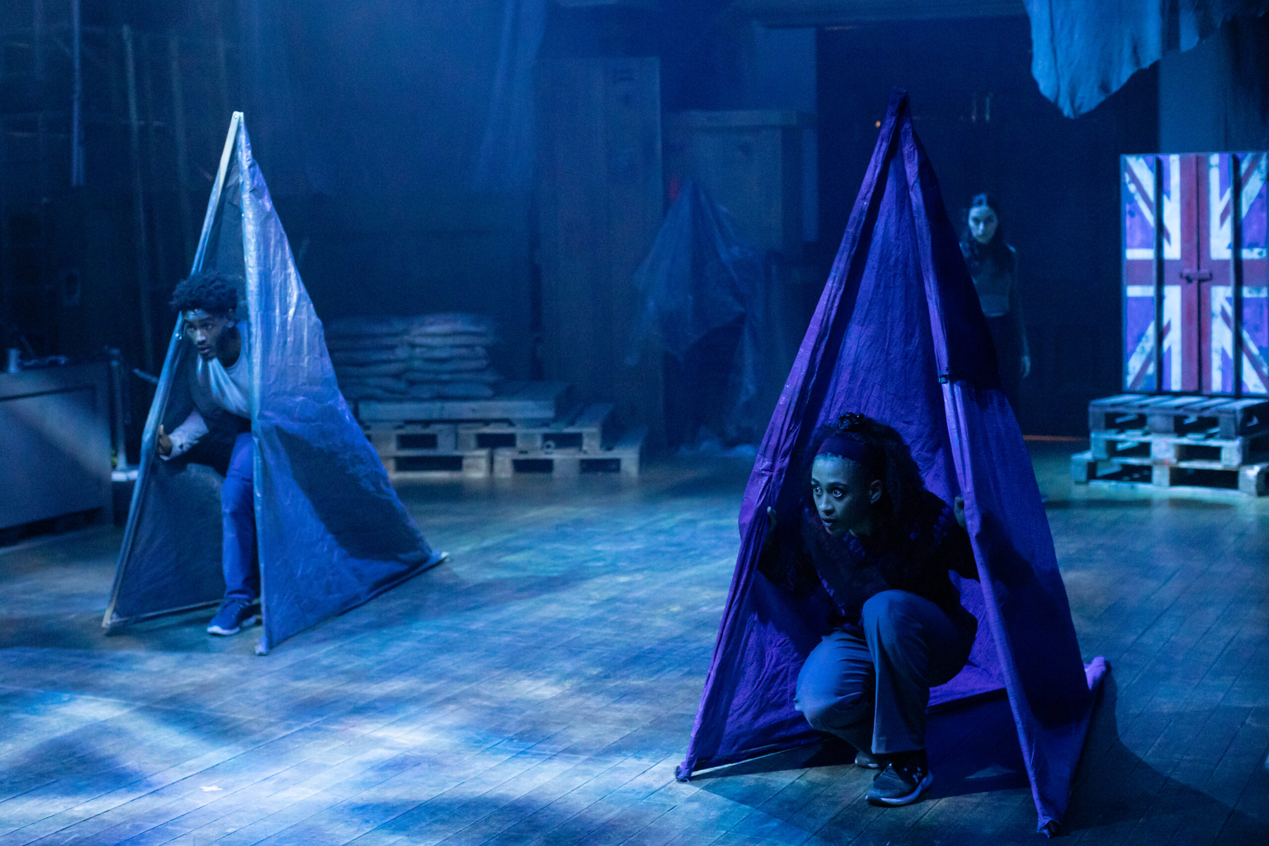 The scene is bathed in a blue light. There are two performers, crouching down inside tent like structures made from blue taurpaulin. in the background you can see a crate with doors painted with a Union Jack.