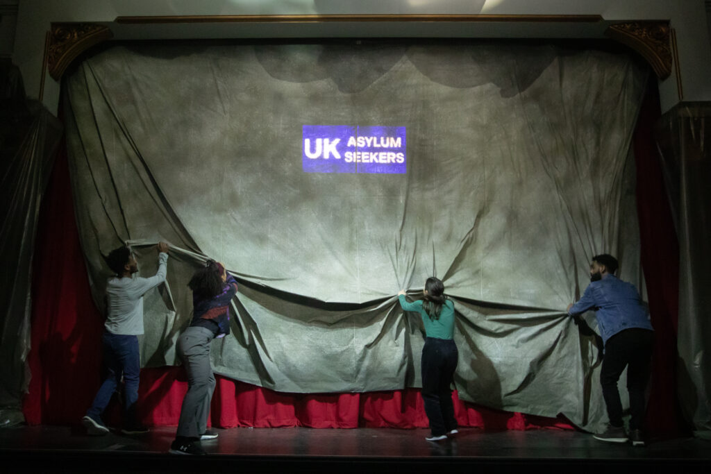 Four performers stand with their backs to the audience, tugging on a large grey curtain as if to pull it down. You can just see the words UK asylum system shining through behind the grey curtain and at the bottom of the curtain you can also see a little bit of red fabric