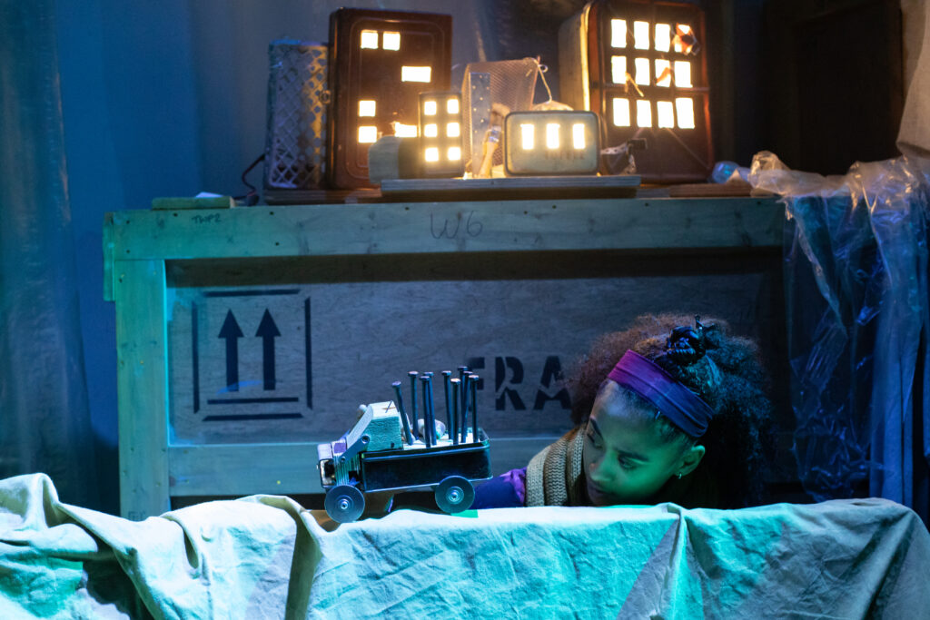 On top of a large wooden crate are cardboard boxes that have been made to look like high-rise buildings with cut out windows and lights inside. Below this a performer holds a toy truck which she is moving across a bench drapped in fabric