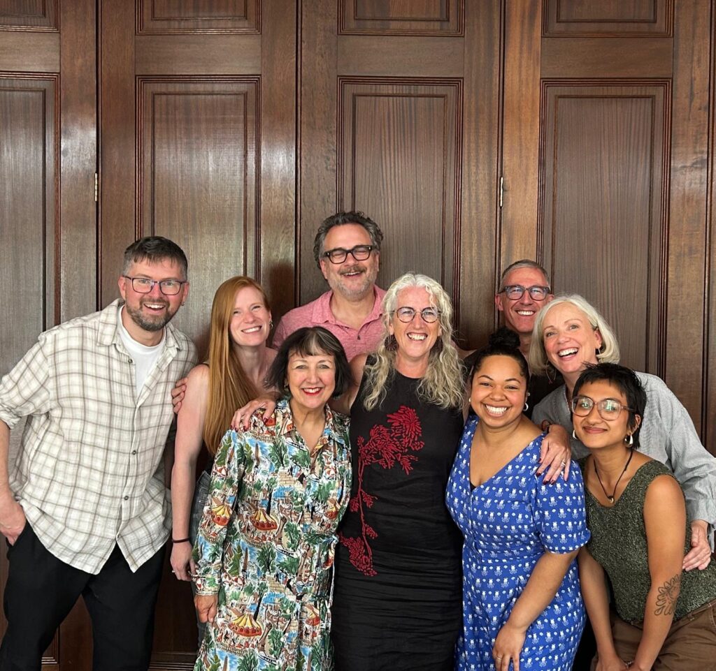 9 people standing in front of wood panelling smiling into the camera