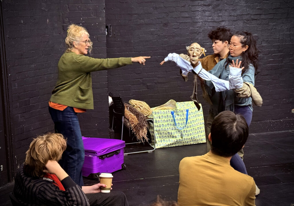 two people with backs to camera watch as a woman in a green cardigan and jeans with white hair tied up in a bun points towards two puppetry students animating a puppet. The puppet wears a shirt with no body but the puppeteers arms inside the sleeves. The puppets head is of an old man with glasses and feathery hair on the side of his head.