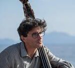 a picture of a man with glasses and a thick head of dark hair sitting outside with his arm draped around a cello