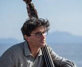 a picture of a man with glasses and a thick head of dark hair sitting outside with his arm draped around a cello