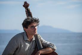 a picture of a man with glasses and a thick head of dark hair sitting outside with his arm draped around a cello