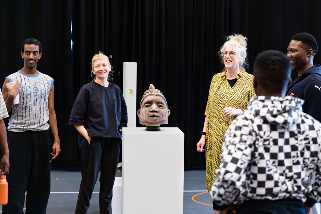 dancers and a puppeteer stand smiling in the rehearsal room with the director and choregorapher. In the middle of the room is a gelede mask sitting on a museum plinth