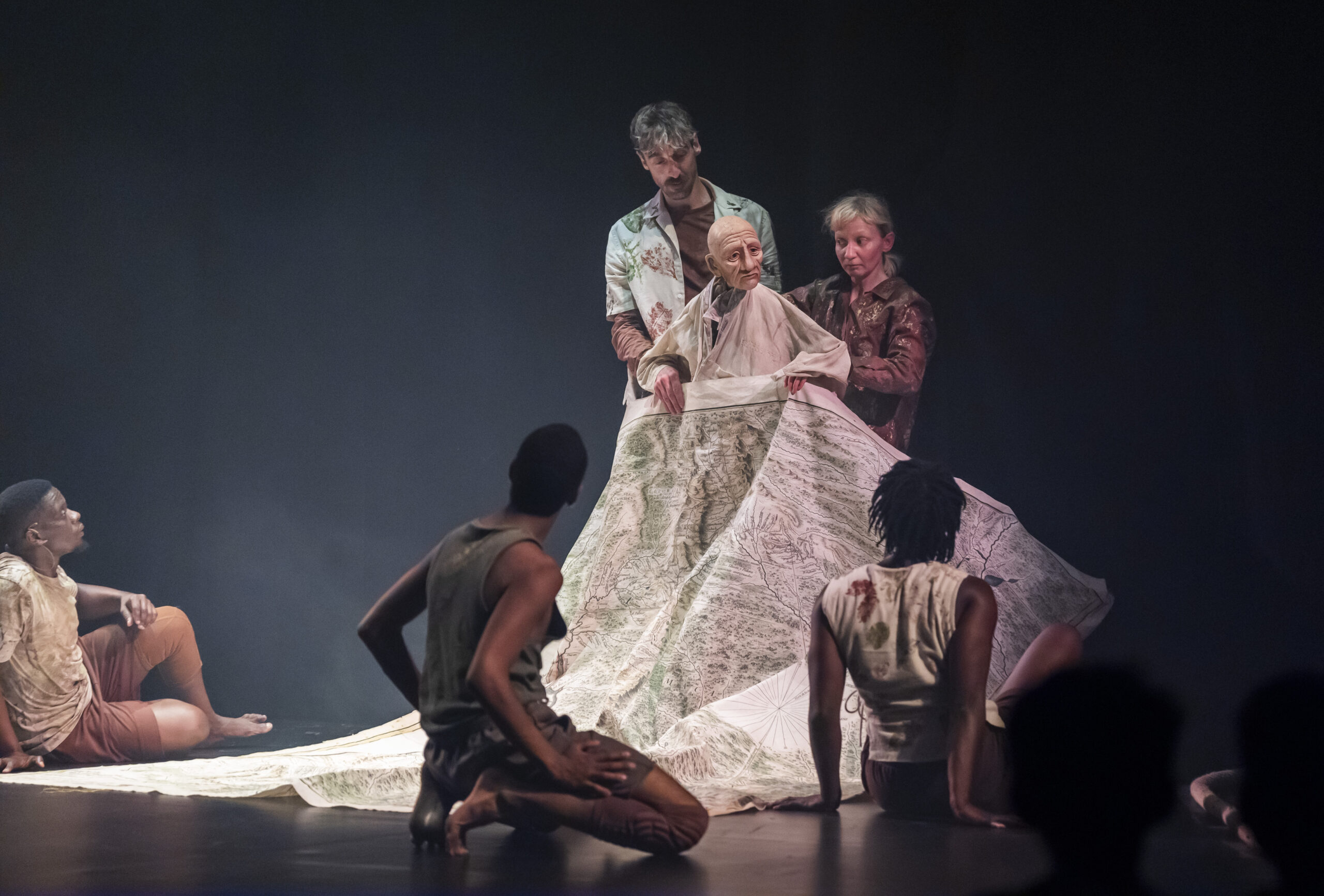 Two puppeteers in floral and earth-toned costumes operate a large, realistic elderly figure puppet draped in a flowing fabric printed with a detailed map, surrounded by performers seated and kneeling on the floor.