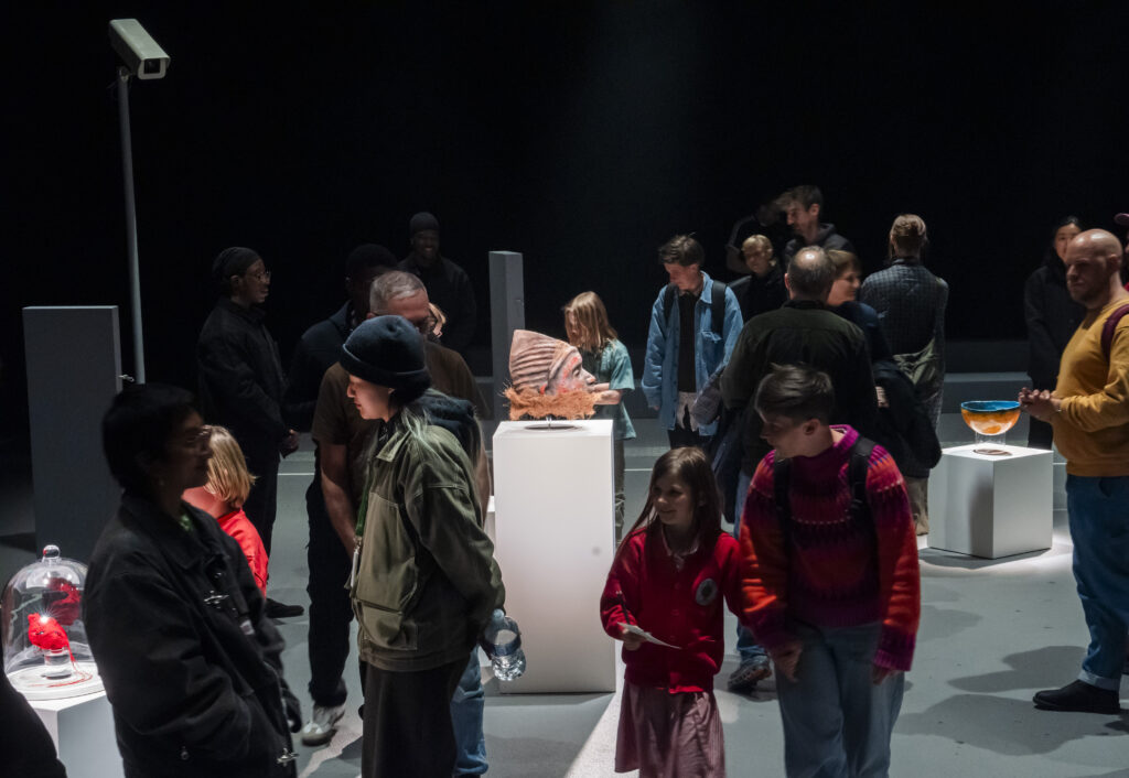 A diverse crowd of audience members browse an exhibition in a darkened space, gathering around illuminated plinths displaying sculptural objects including a mask-like Gelede head and a glowing red heart under a glass dome.