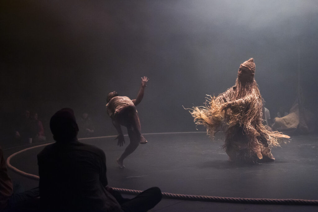 A dancer in a simple costume bends backwards on a circular stage opposite a towering figure entirely covered in a shaggy straw and fibre costume, in misty atmospheric lighting.