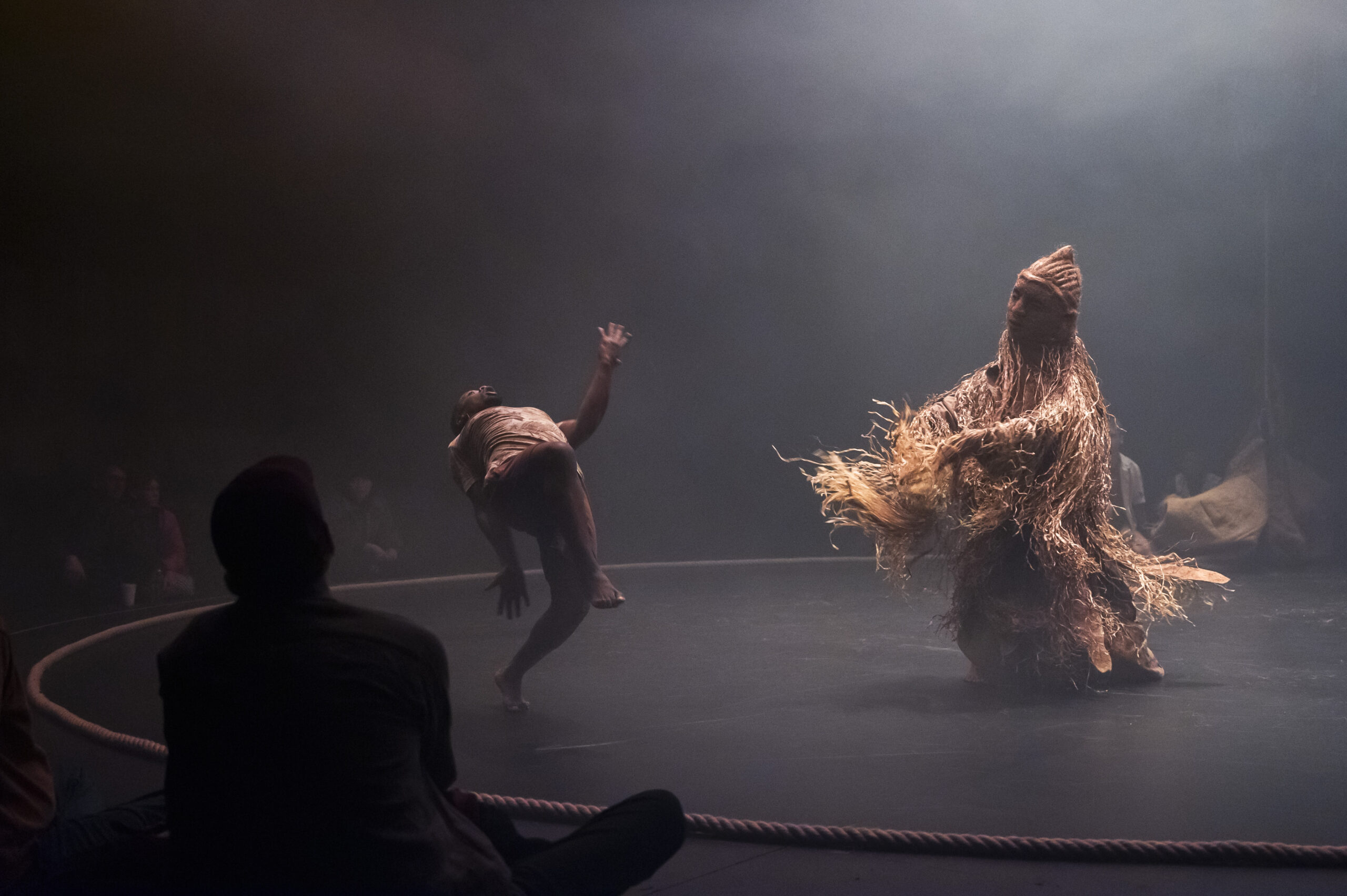 A dancer in a simple costume bends backwards on a circular stage opposite a towering figure entirely covered in a shaggy straw and fibre costume, in misty atmospheric lighting.