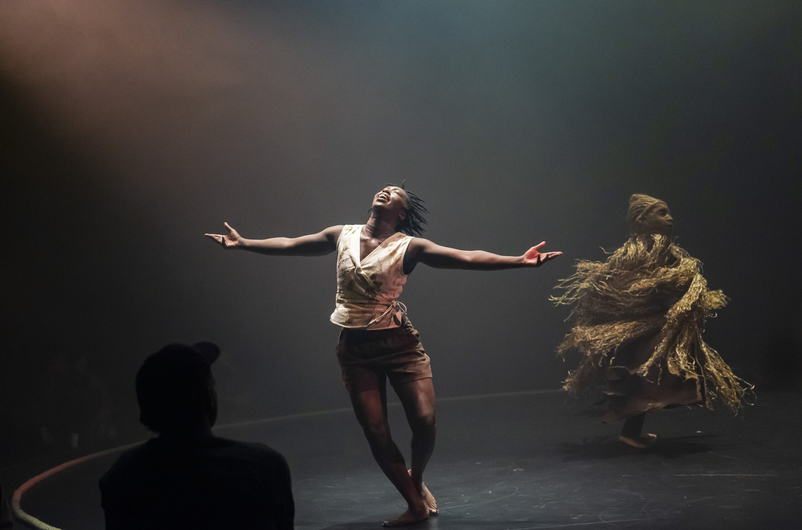 A barefoot performer in a cream wrap top and brown shorts leans back with arms outstretched and head raised, mid-dance. In the background, another figure moves in a voluminous straw-like costume.