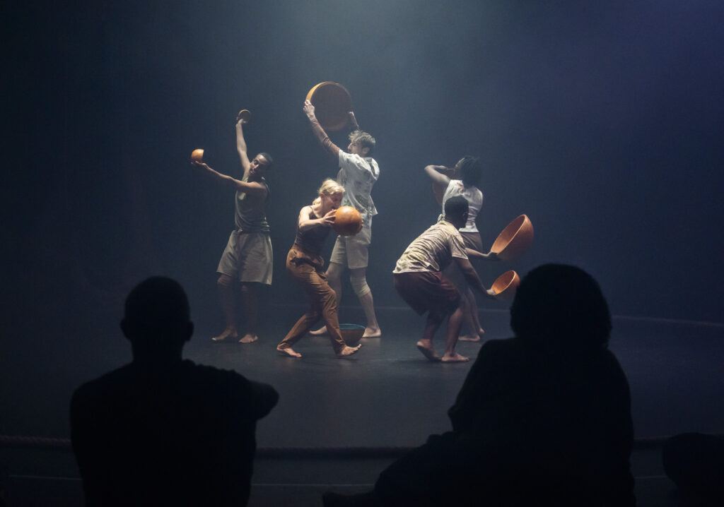 Five performers in earthy, minimal costumes handle large curved wooden bowls on a dimly lit stage, silhouetted audience members visible in the foreground.