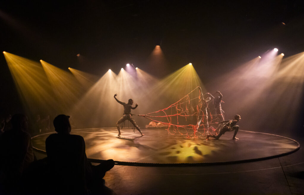 Three performers in patterned bodysuits manipulate a large red rope net on a circular lit stage, surrounded by atmospheric yellow and white stage lighting and a haze of smoke.