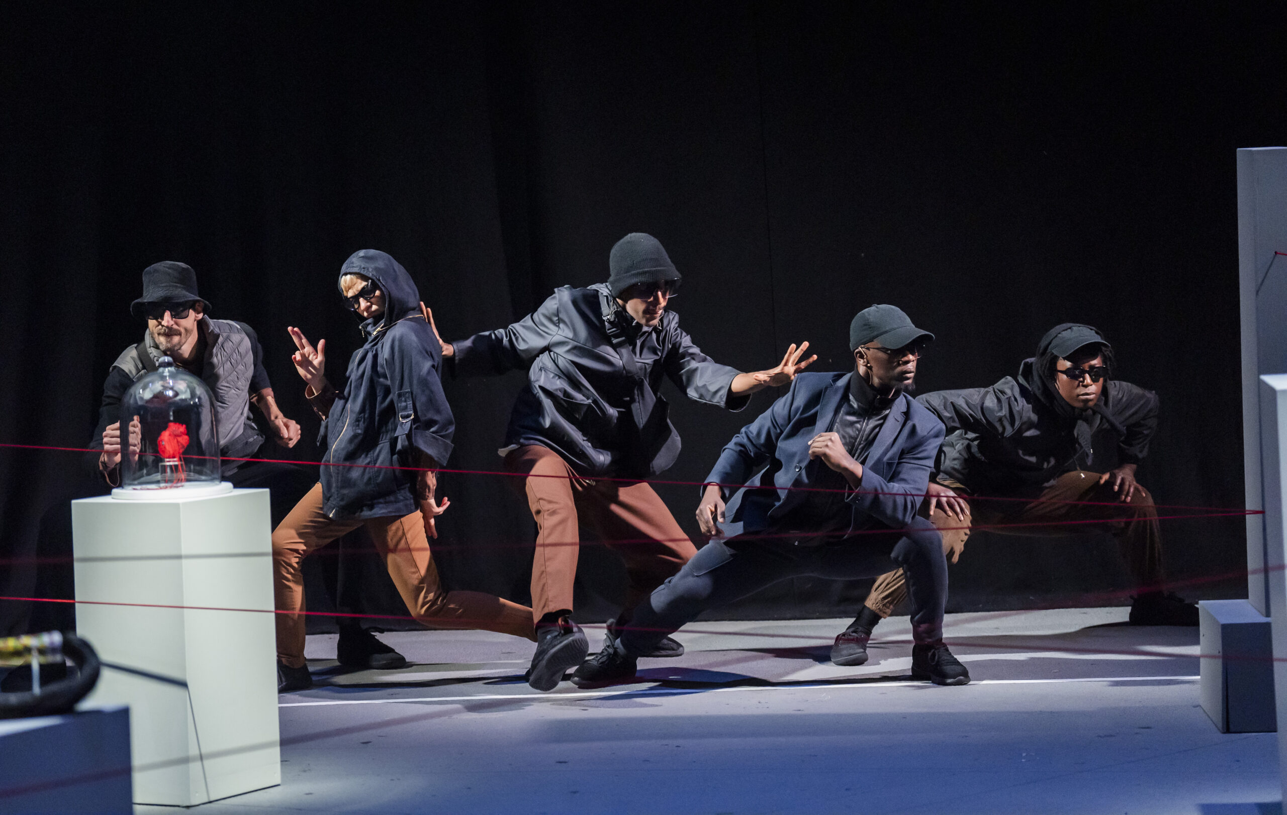 Five performers dressed as thieves in dark hooded jackets, caps, and sunglasses crouch in choreographed poses on a pale stage, red string criss-crossing in front of them. A red sculptural heart sits on a plinth to the left.