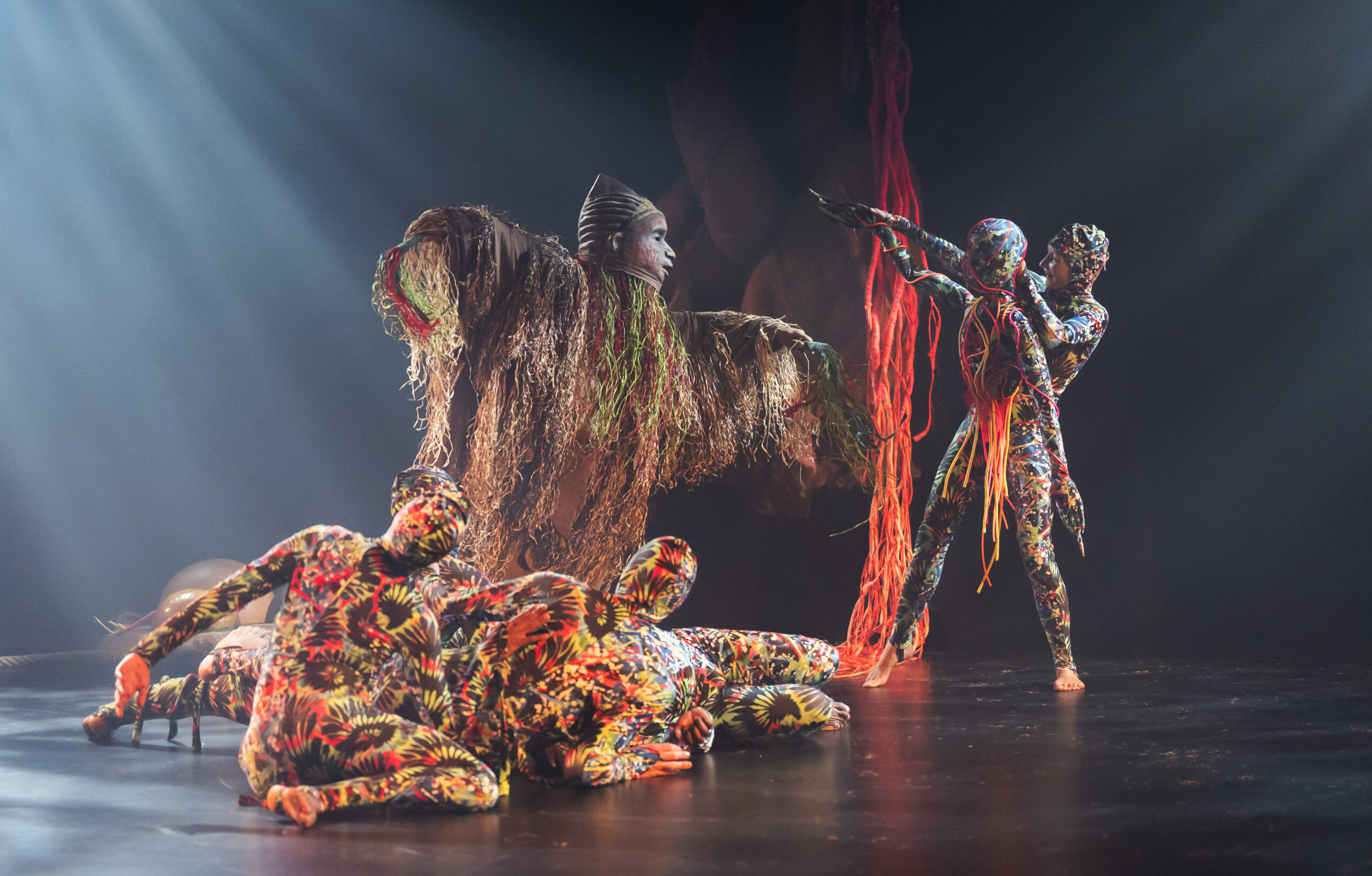A large figure in an elaborate raffia and natural-fibre mask costume stands centre stage surrounded by performers in vivid, colourful floral bodysuits, with cascading red and orange rope hanging from above.