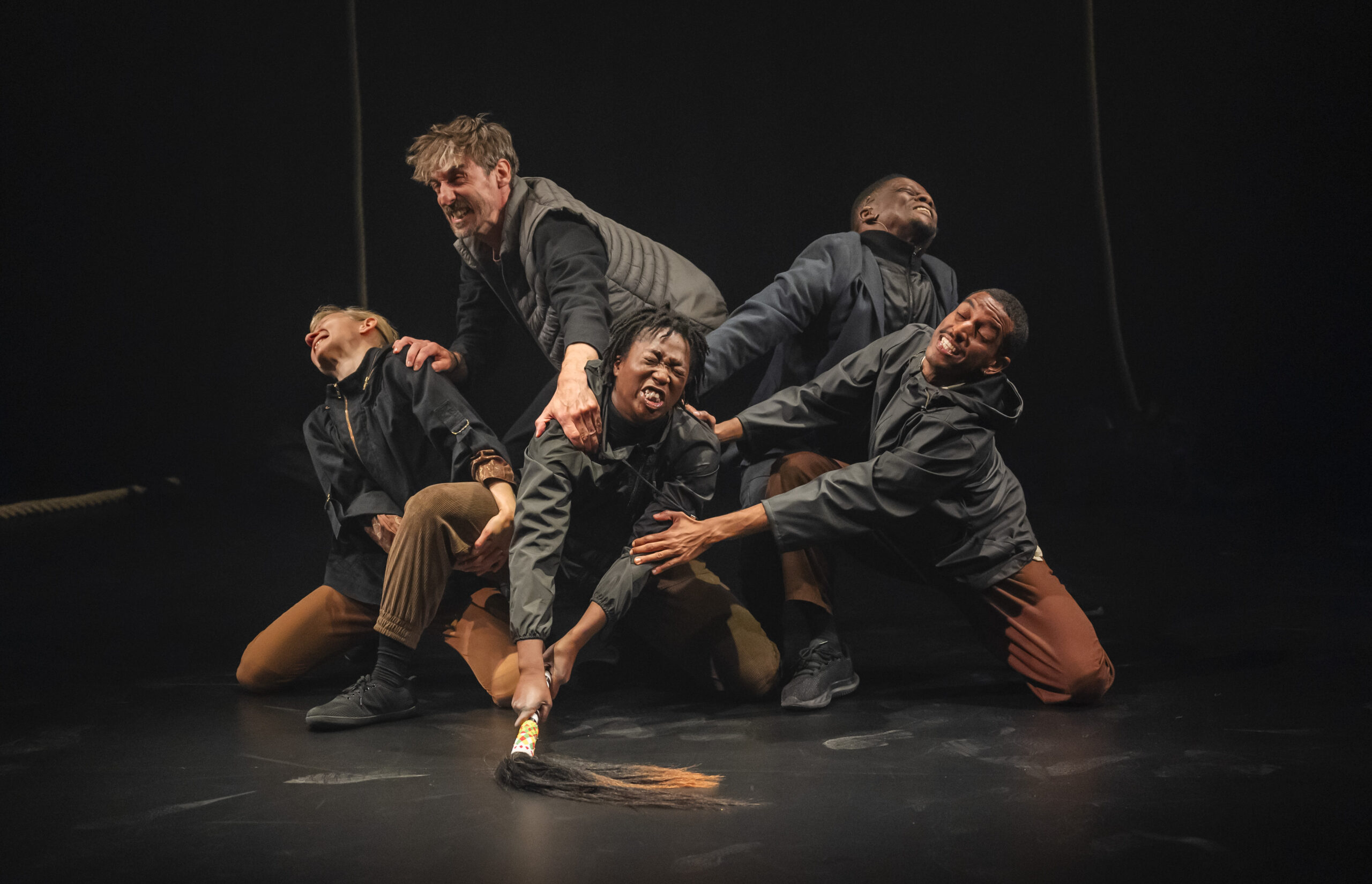 Five performers in a tangled, dynamic group on a black stage floor, bodies intertwined in an intense physical theatre tableau. One performer at the centre appears to be struggling to pull a stick, with long black hair attached to the end, from the ground. The other four performerr around her are joined in the struggle to pull the object.