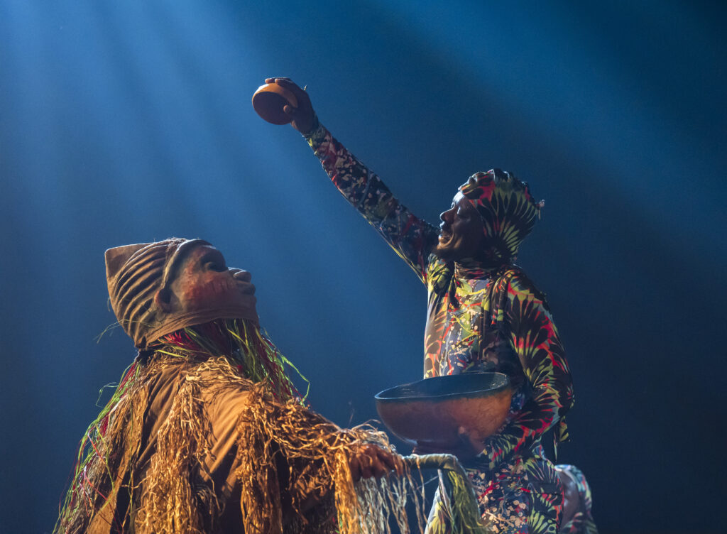 Two richly costumed performers — one in a vivid floral-print bodysuit, one in a raffia and natural fibre costume — reach upward holding clay bowls, lit dramatically in deep blue light.