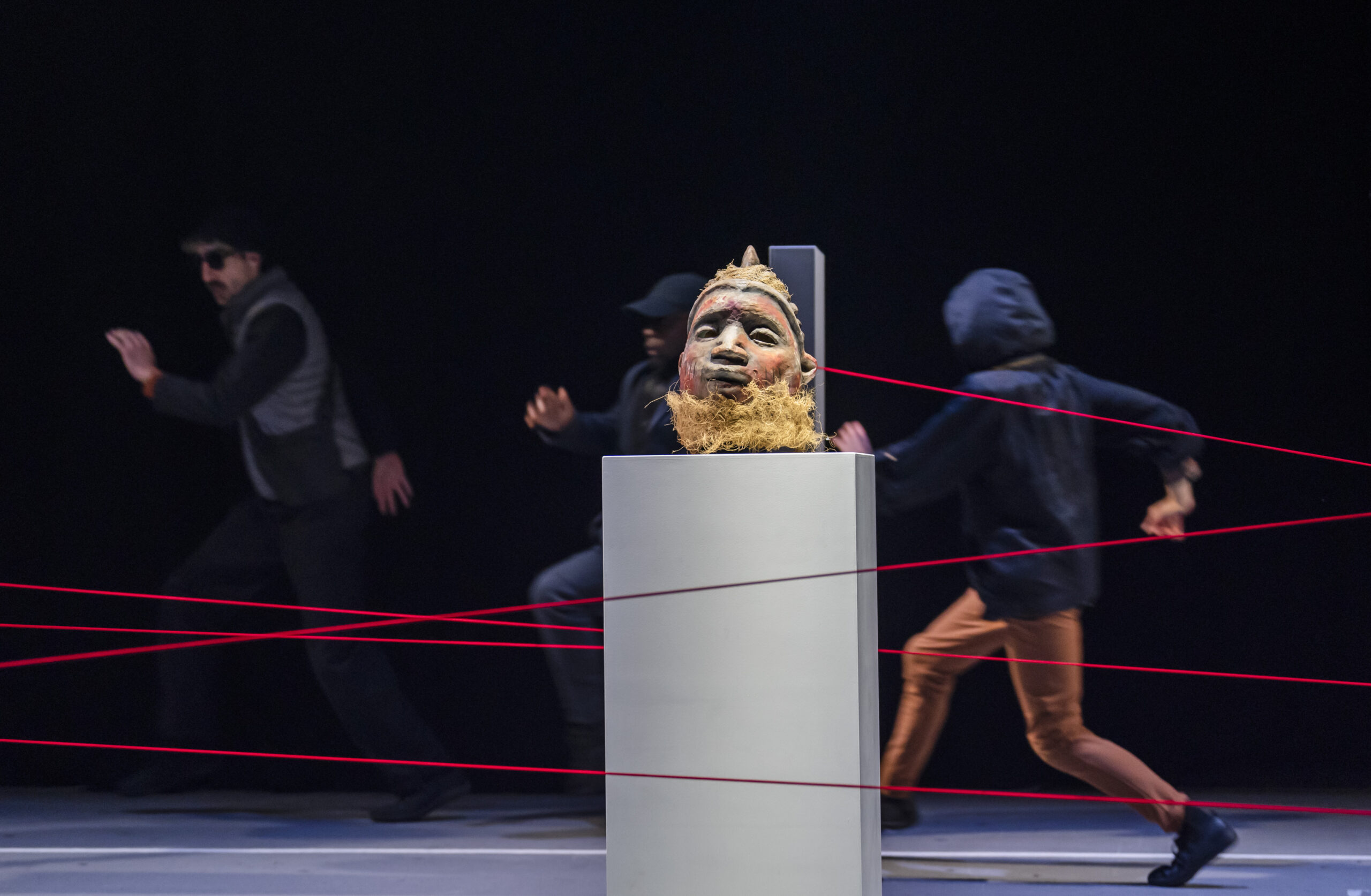 A traditional carved African Gelede mask rests on a white museum plinth in sharp focus, while figures in dark clothing run blurred through a web of criss-crossing red strings behind it.