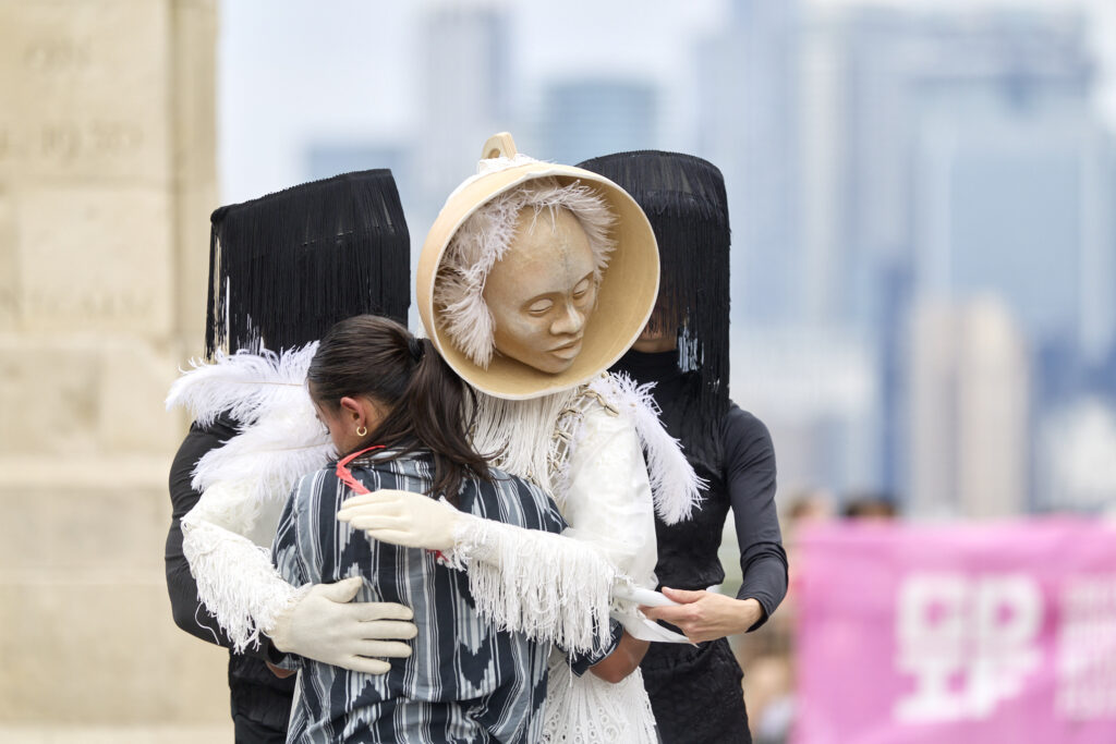 A dancer with long ponytail and her back to the camera, hugs a Old Spirit puppet dressed all in white with feathers and a face made out of wood. Two puppeteers dressed in black animate the puppet.