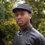a young black man with a moustache, earrings and wearing a black cap and black leather jacket with a large necklace over the jacket.