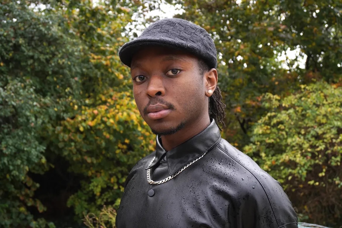 a young black man with a moustache, earrings and wearing a black cap and black leather jacket with a large necklace over the jacket.