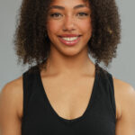 A smiling black woman looking straight to camera, with long curly hair, wearing a black vest top and a nose ring.