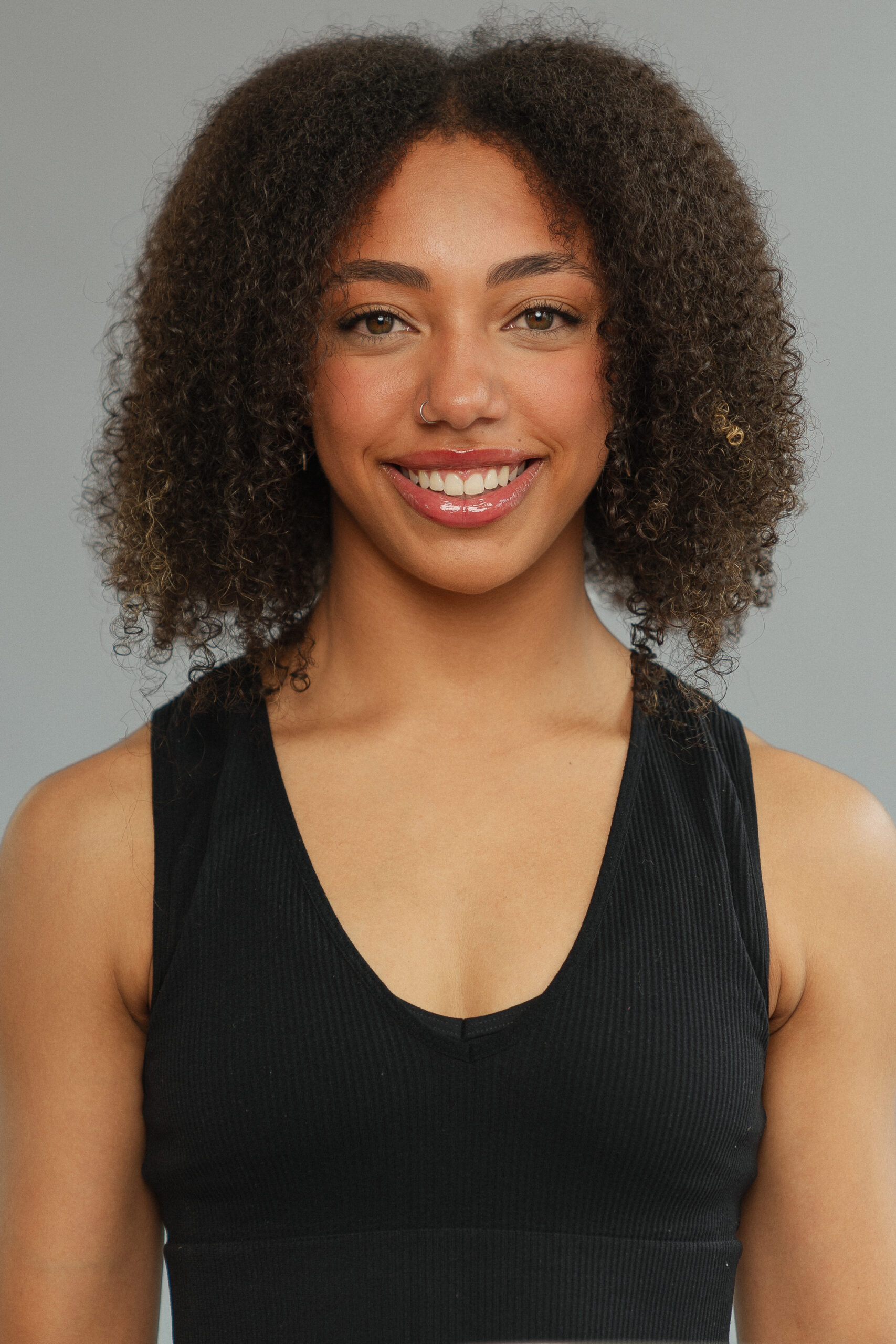 A smiling black woman looking straight to camera, with long curly hair, wearing a black vest top and a nose ring.