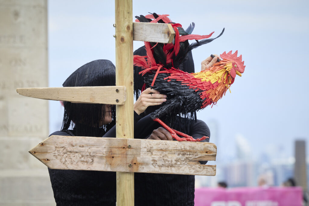 a red, yellow and black puppet of a Rooster is animated by two puppeteers dressed in black. The Rooster is perched on a crossroads sign