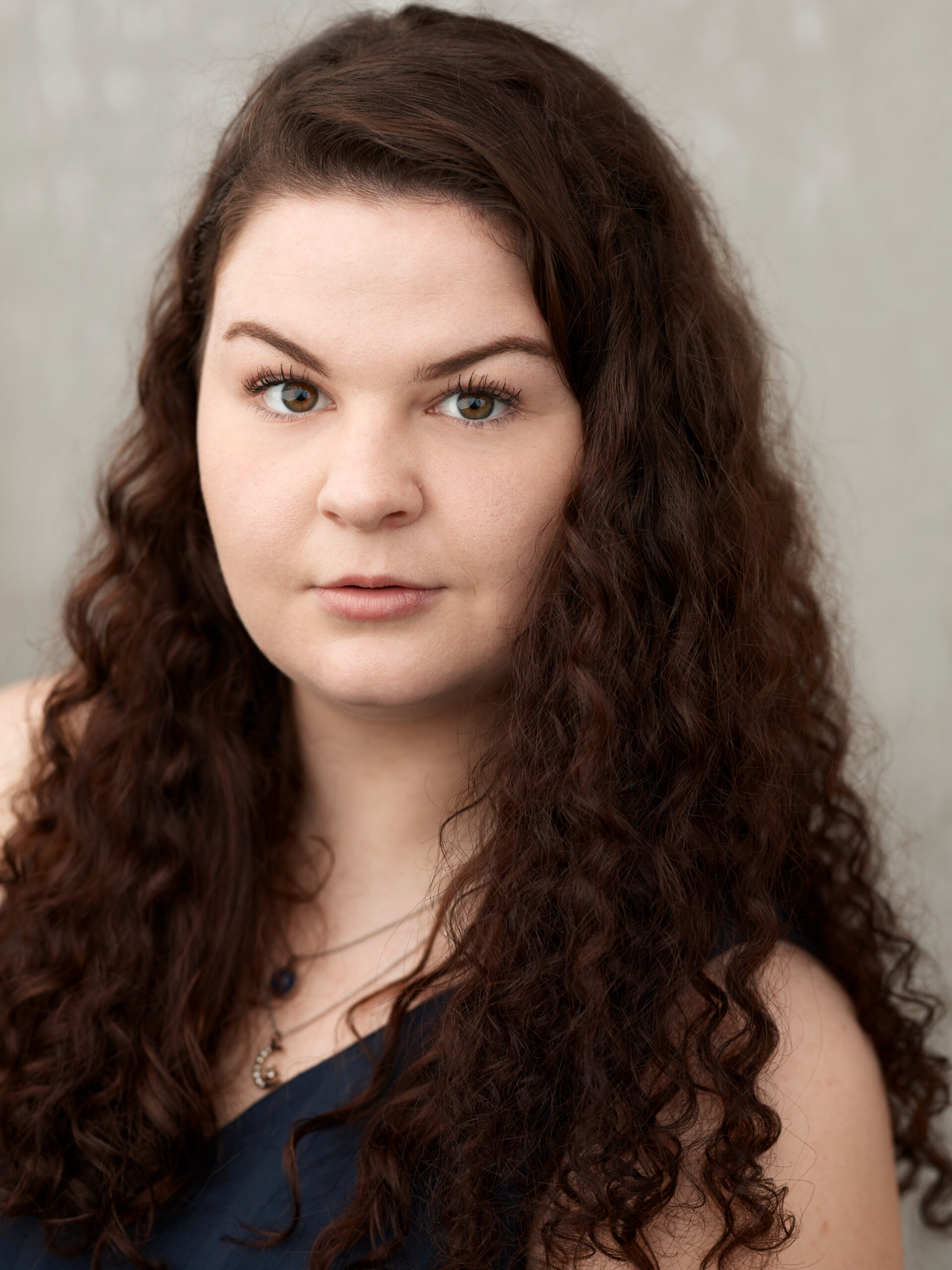 Professional headshot of a woman with long, curly brown hair against a neutral gray background. She has fair skin, hazel eyes, and light makeup, and wears a dark navy top with a delicate layered necklace. Her expression is calm and direct, looking straight at the camera.